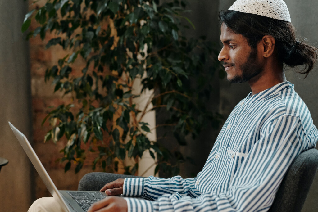 Person with dark skin tone smiles into laptop. They are seated on a chair with houseplants in the background.