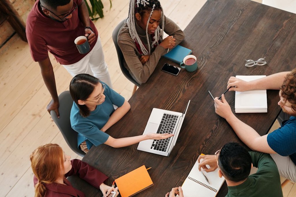 Group of diverse persons gather around a wooden table with their laptops.