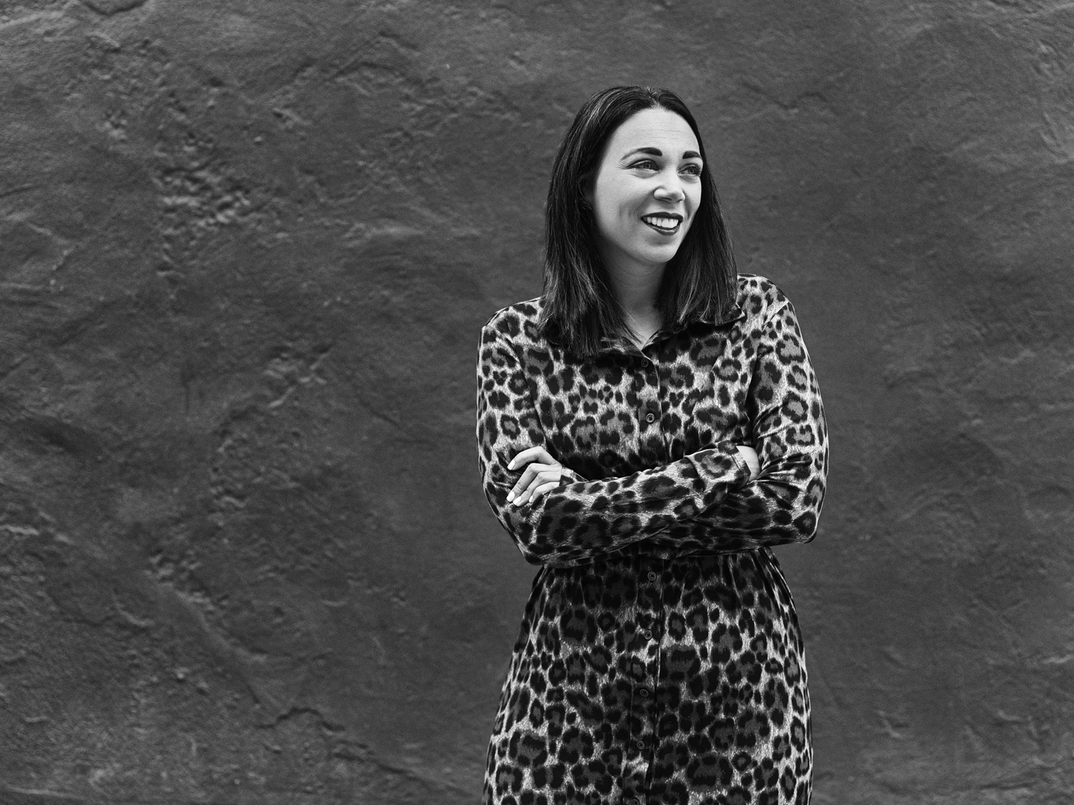 Black and white photo of Shauna Cole. She is wearing an animal print dress and standing against a cement wall.
