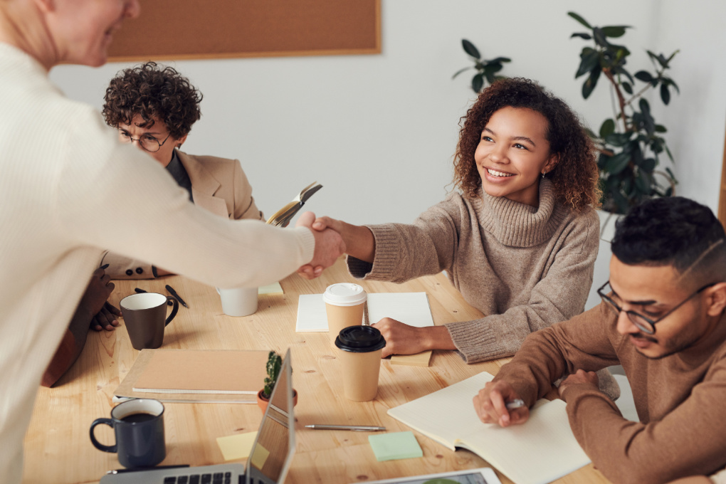 Diverse group of employees gather around a table. One is shaking hands with another.